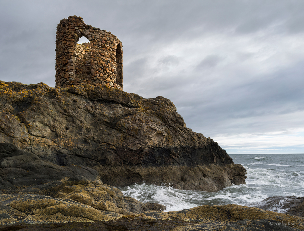 An image of the Lady's Tower in Elie. The choppy sea water is crashing against the rocks under the Lady's Tower turret. A stormy sky is in the background.