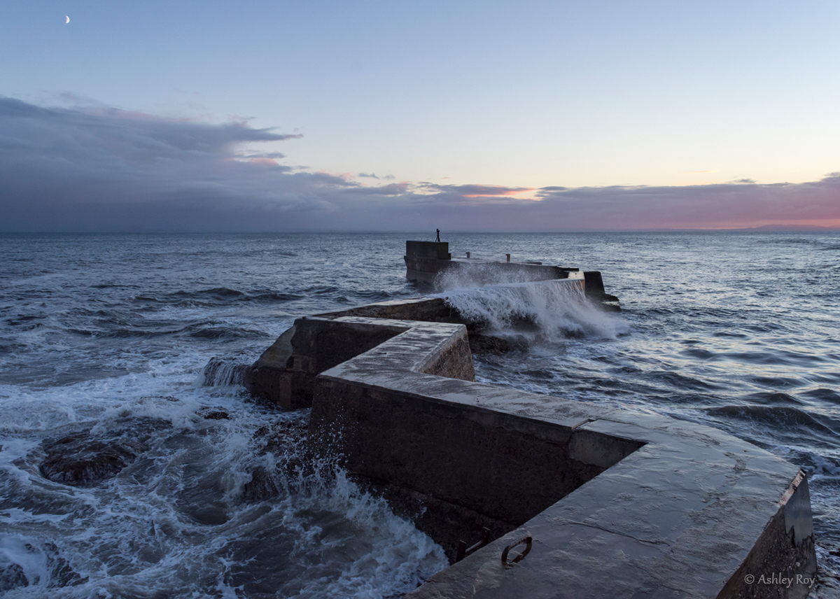A landscape image of the breakwater at St. Monan's Harbour. It is dusk, the sky is pink and blue, and the seawater is crashing over the jagged breakwater.