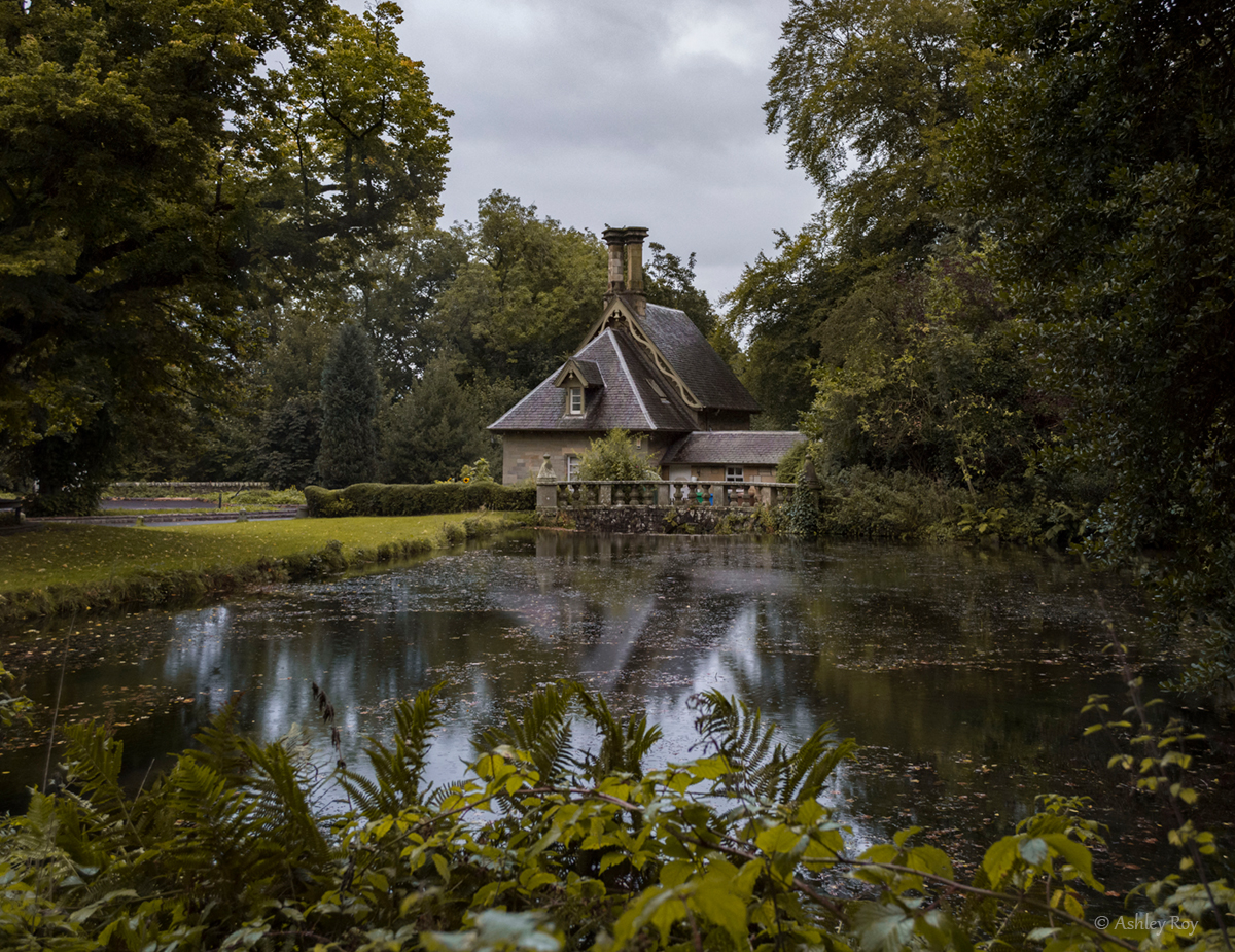 An image of the gatehouse at Falkland Estate. The gatehouse is in the background, enclosed between large trees with a rippled pond in the foreground.