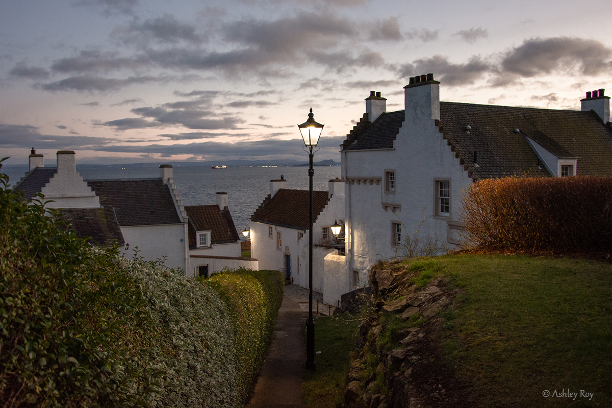 A landscape image of the Pan Ha pathway in Dysart. The sea is in the background, with the Pan Ha houses on either side of the pathway. The sky is setting and stormy.