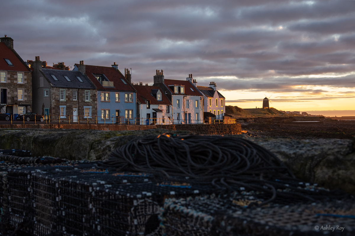 A landscape image of the houses along St. Monans Harbour. It is dawn, and the morning light is reflected on the front of the houses. A windmill is in the distance, and creels are stacked up in the foreground.
